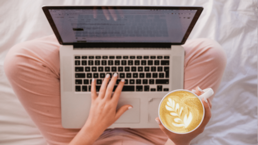 A woman with white painted fingernails sits on a duvet crossed legged. Her laptop is resting on her knees, while one hand types on her MacBook and the other holds a cup of Latte style coffee.