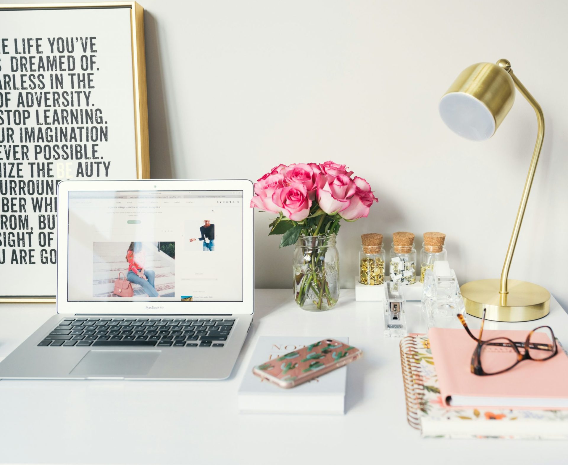 A photo of a clean white desk with a laptop sat on top of it. To the right side are some pink flowers in a small vase and a golden table lamp.
