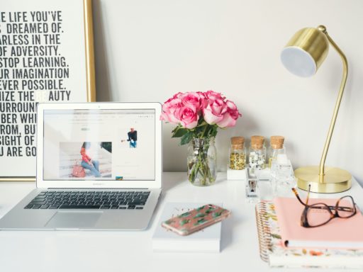 A photo of a clean white desk with a laptop sat on top of it. To the right side are some pink flowers in a small vase and a golden table lamp.