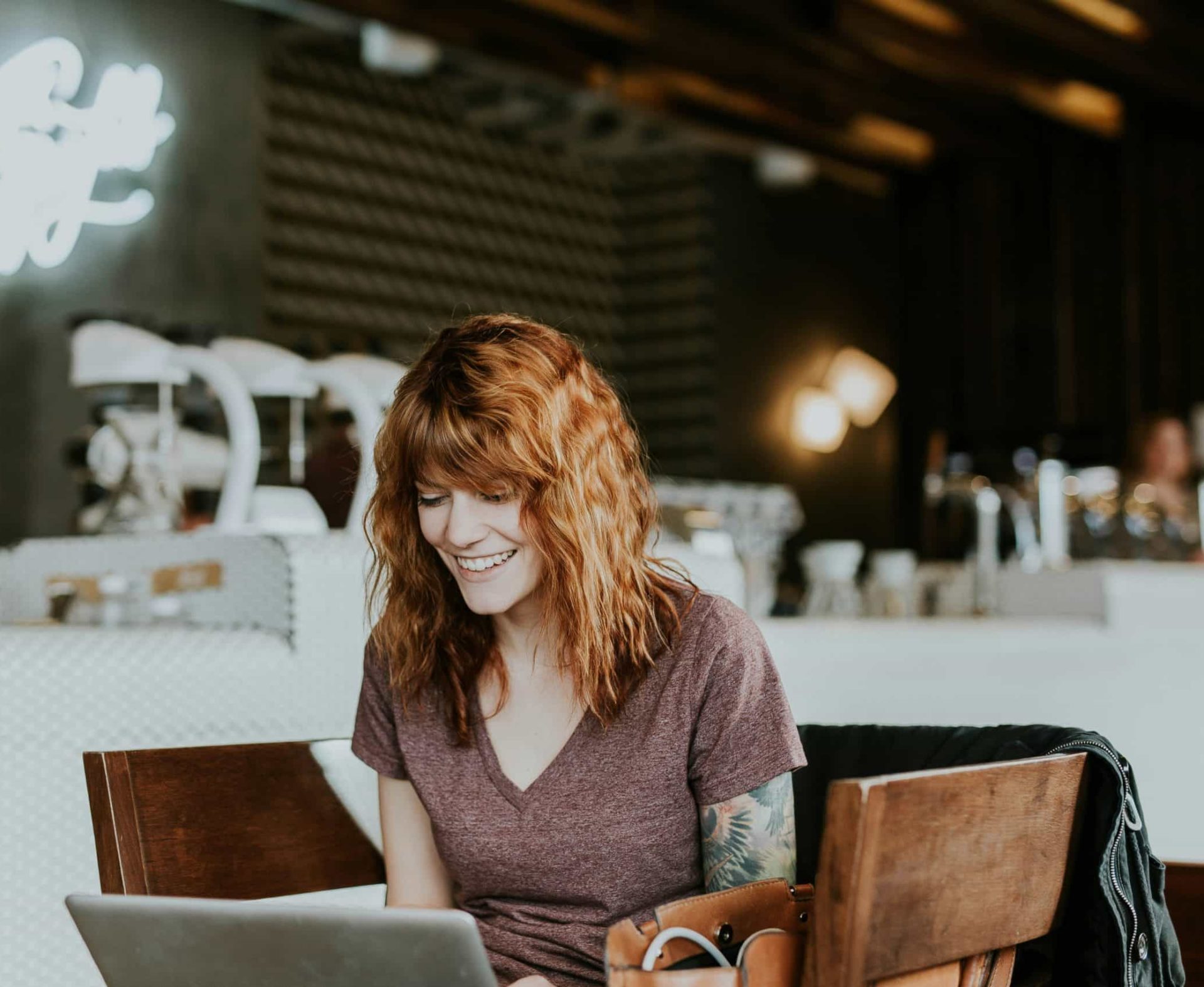 Ginger-haired woman smiles at her laptop screen while sat in a hipster coworking space. Presumably so overjoyed because she's just found out how amazing it is using Quuu for social media content suggestions using AI.